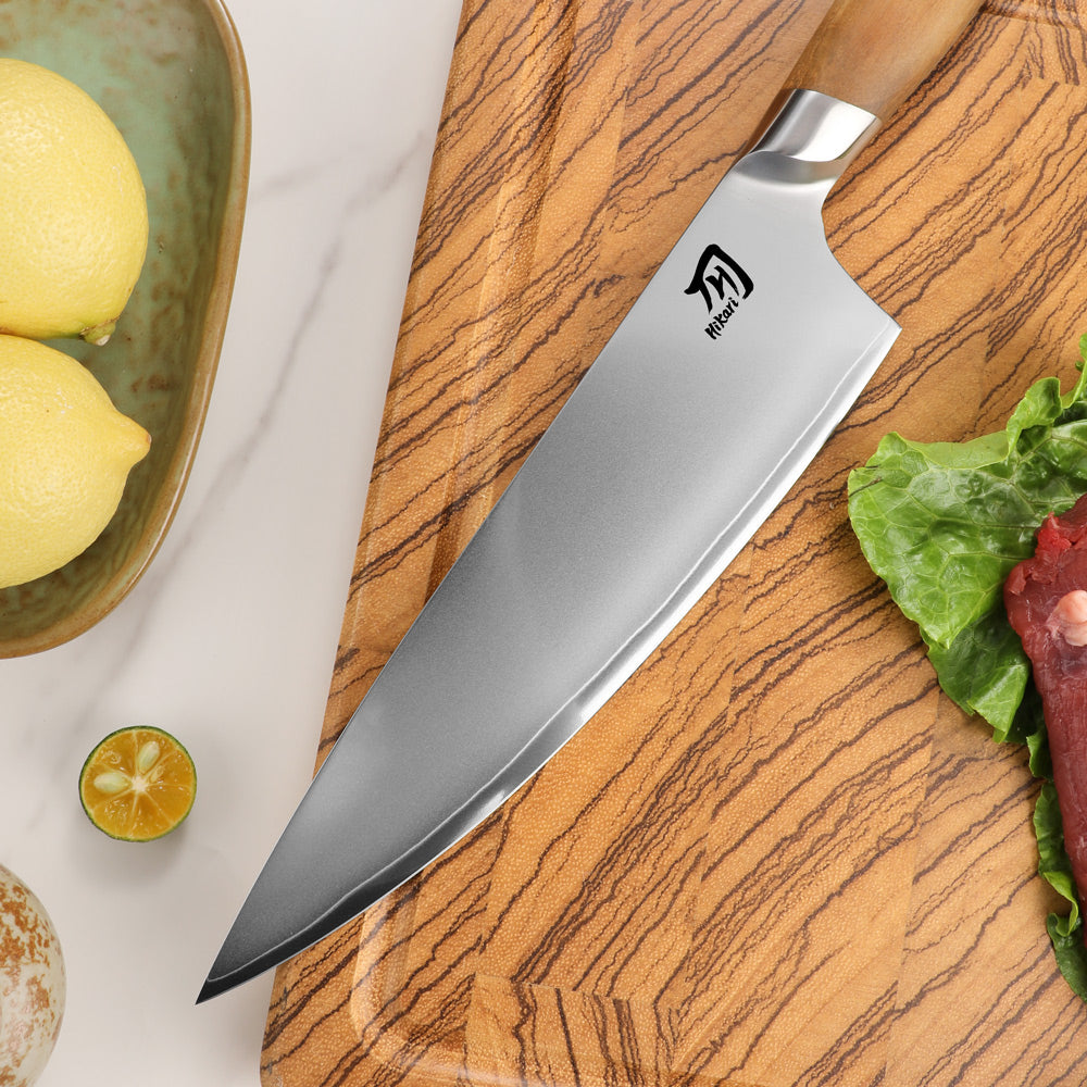 A chef's knife with a silver blade and light wood handle resting diagonally on a wooden cutting board, surrounded by kitchen props including lemons, lettuce, and raw meat.