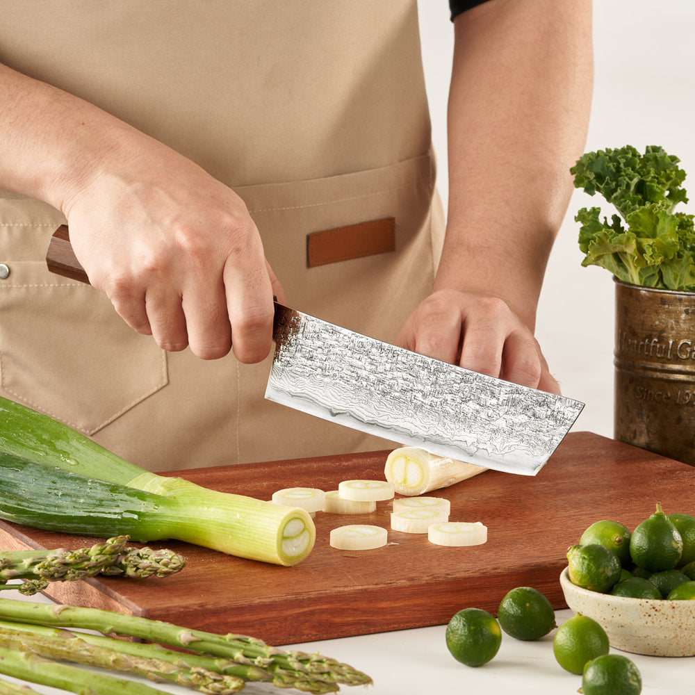 Person cutting vegetables on a wooden board with a chef's knife, surrounded by fresh produce.