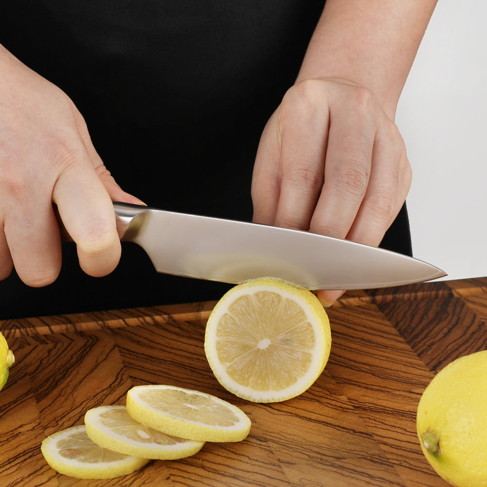 A close-up of a person's hands slicing a lemon with a stainless steel chef's knife on a wooden cutting board, with lemon slices scattered around.