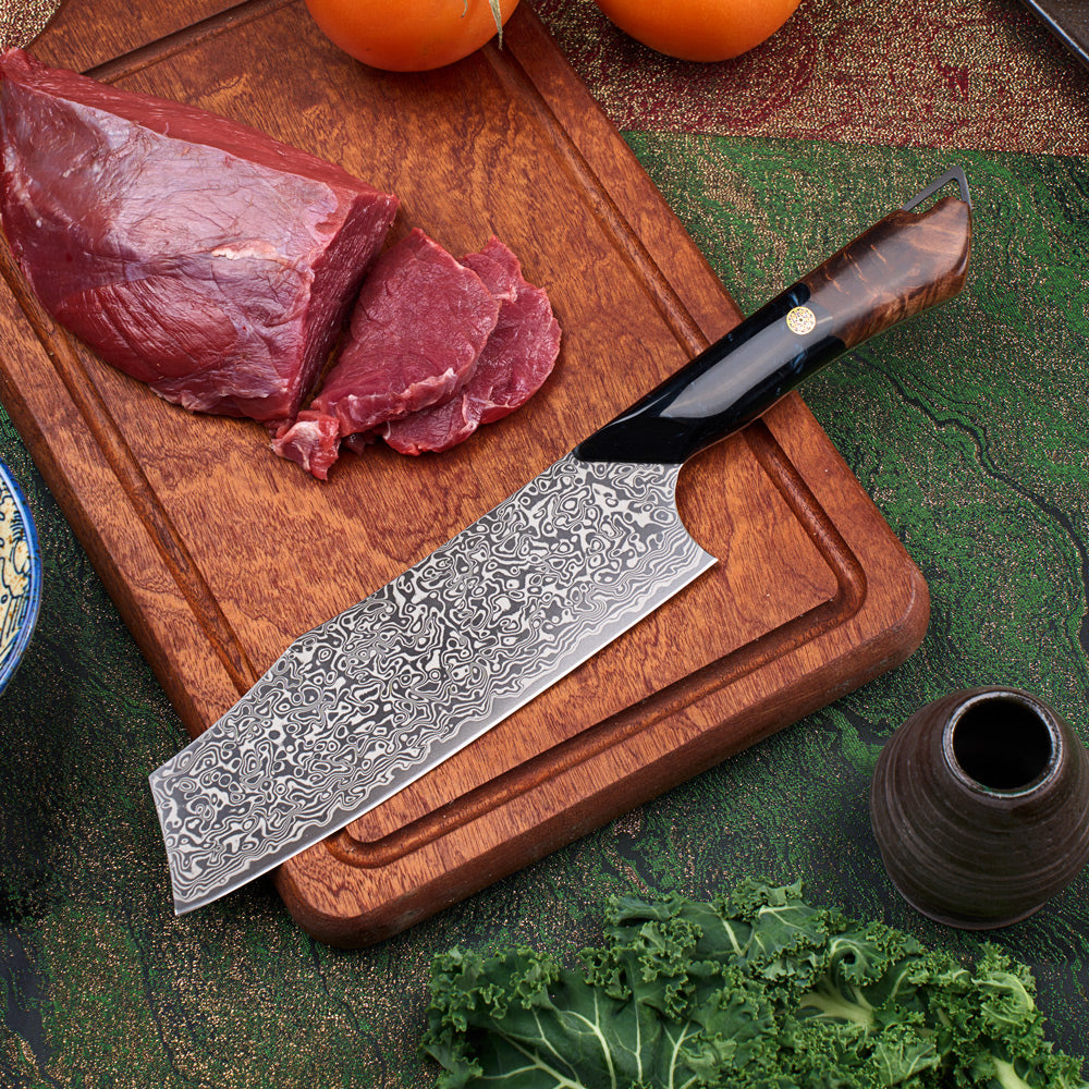 A Damascus steel kitchen knife with a dark wood handle rests on a wooden cutting board next to raw beef and sliced meat, with kale and a small vase in the foreground.