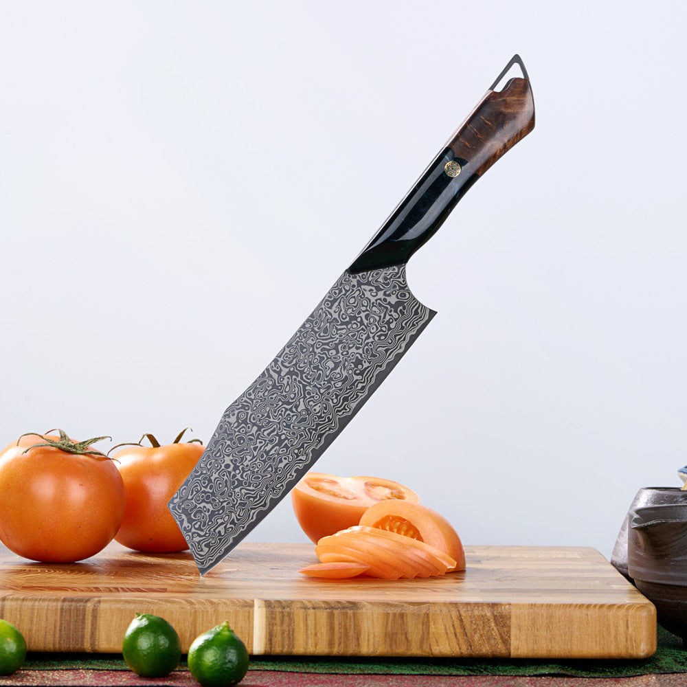 A chef's knife with a patterned Damascus steel blade and a dark wood handle is shown standing upright on a wooden cutting board, with tomatoes, limes, and sliced tomato around it on a light gray background.